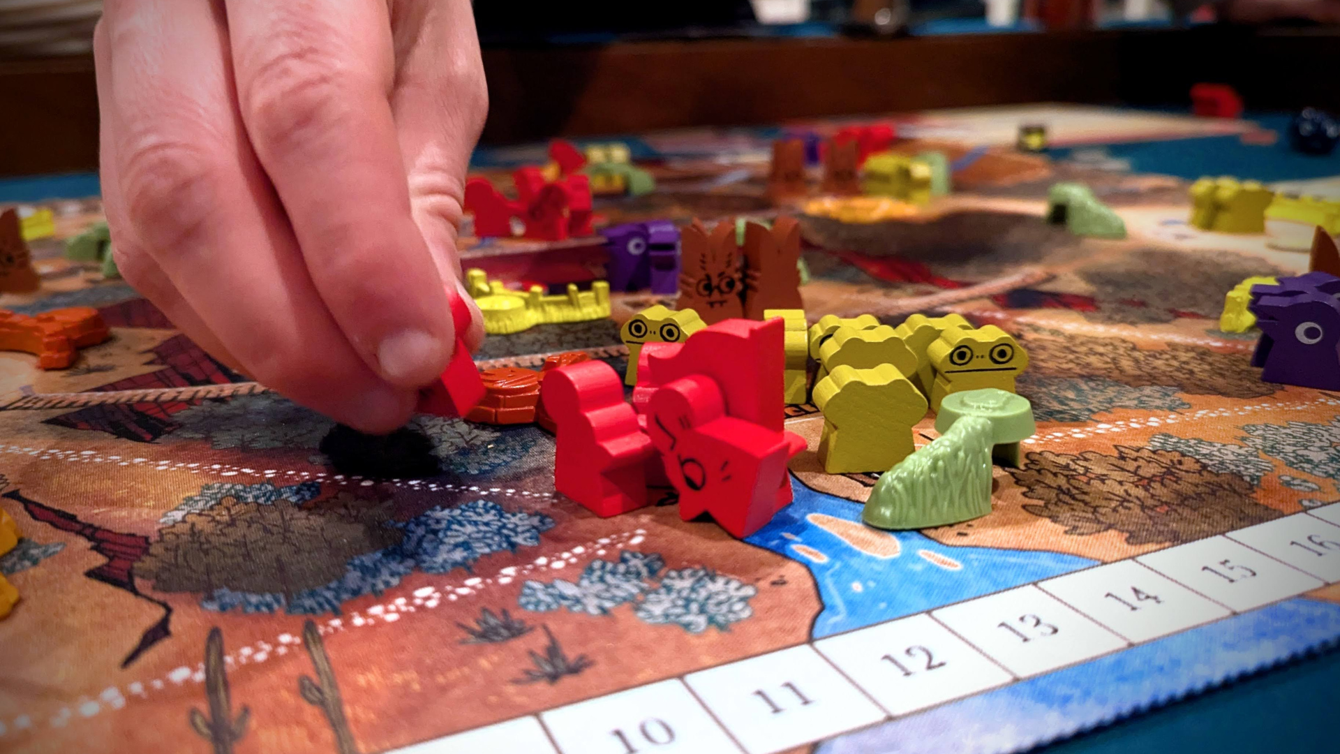 A hand moves a red wooden game piece on a colorful board game featuring various animal-shaped pieces, reminiscent of the whimsy in "Meet the Member: With Flour in Her Hair," with a numbered score track along the edge.