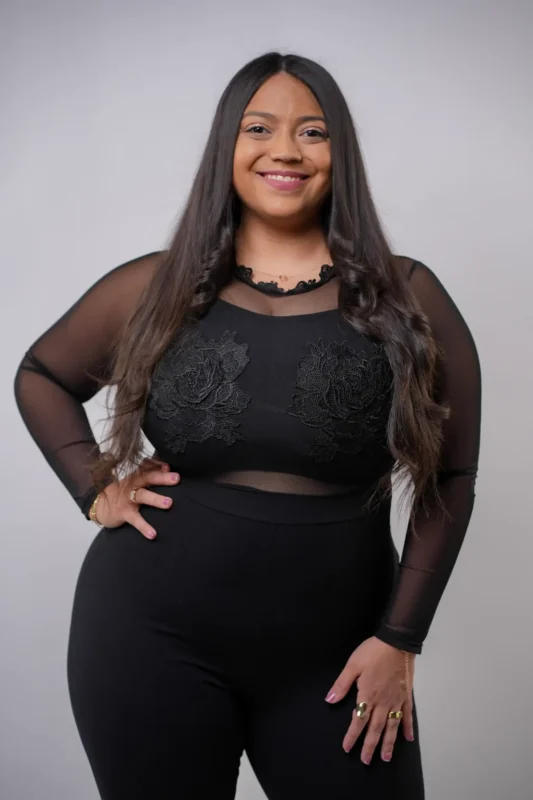 A woman with long dark hair stands against a plain light gray background. She is wearing a black outfit with sheer sleeves and floral patterns on the top. She smiles at the camera with one hand on her hip.