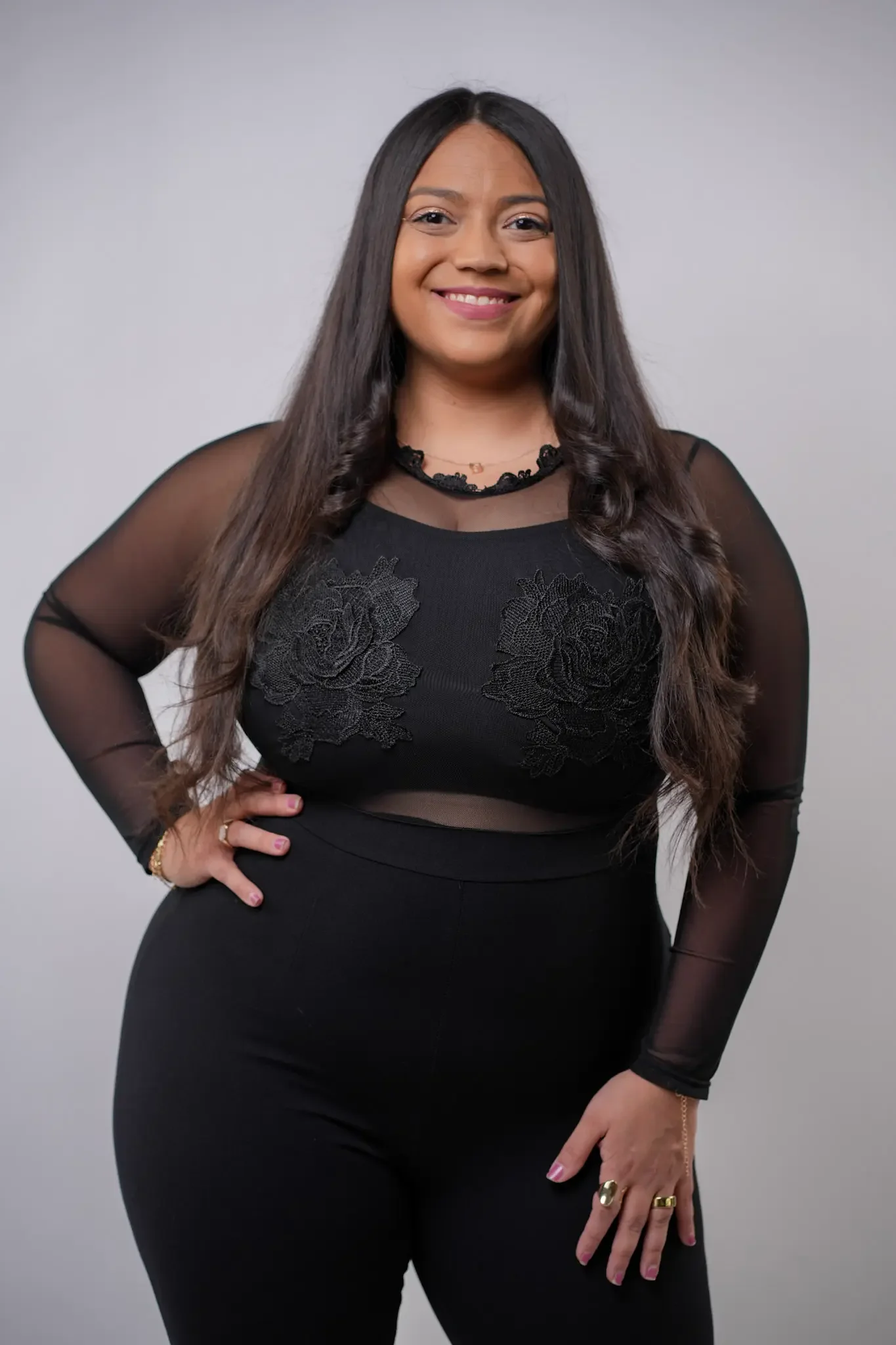 A woman with long dark hair stands against a plain light gray background. She is wearing a black outfit with sheer sleeves and floral patterns on the top. She smiles at the camera with one hand on her hip.