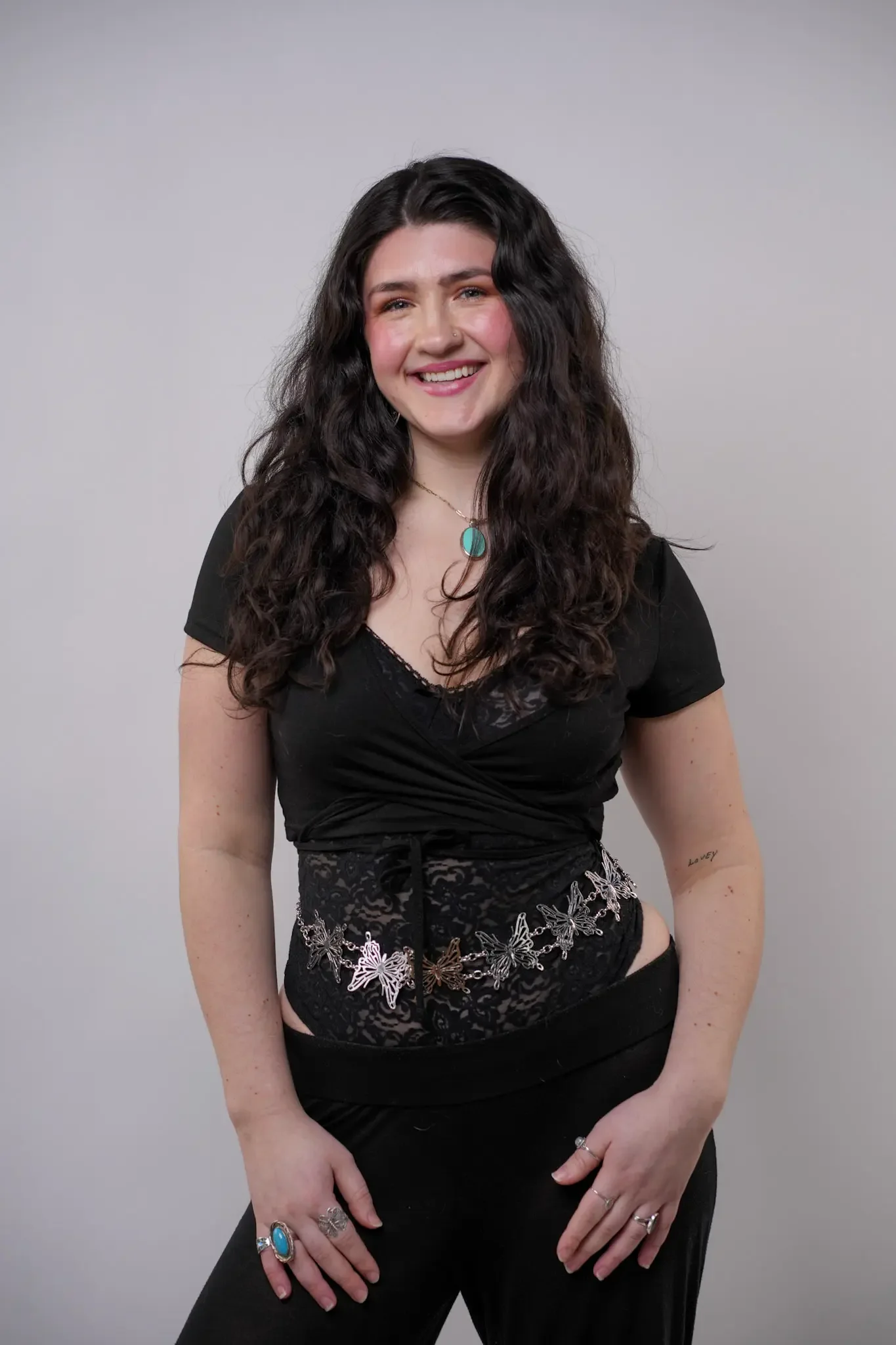 A woman with long, wavy dark hair stands against a plain light gray background. She is wearing a black top, black lace corset, black pants, turquoise jewelry, and is smiling at the camera.