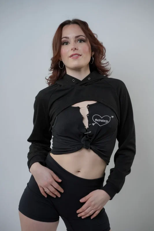 A person with wavy brown hair stands against a plain background, wearing a black hoodie tied at the waist over a ripped black t-shirt with a heart graphic, and black shorts.