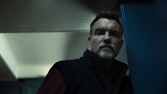 A man with short hair and a beard stands indoors, leaning against a blue wall. The lighting is dim, creating shadows on his face and the background. He has a serious expression and is looking slightly downward.