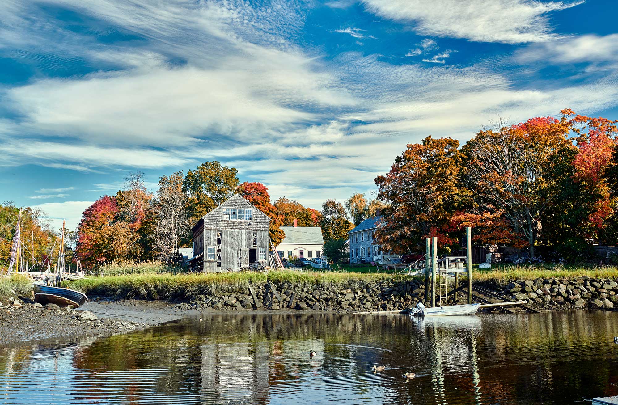 A calm river with a small dock and boats, surrounded by trees with autumn foliage. An old wooden barn and houses rest in the background under a partly cloudy sky, where creativity is liberation, reflected in the tranquil water.