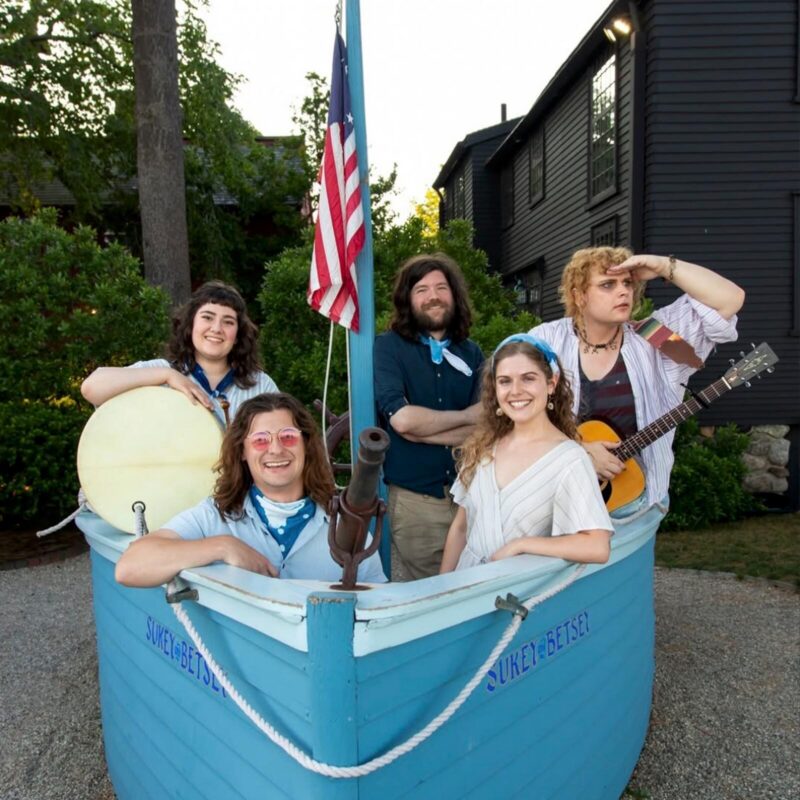 Five people pose in a small blue wooden boat labeled "SUKey BETSEL," with one holding a guitar, another a drum, and an American flag behind them—radiating queer music vibes in their casual, nautical-themed clothing.