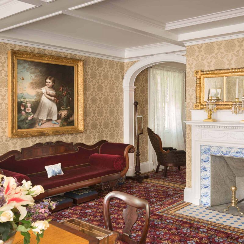A Victorian-style living room with patterned wallpaper, a red velvet sofa, a large framed portrait of a child, a white fireplace with blue tile accents, and a mirror above the mantel. Flowers sit on a wooden table.