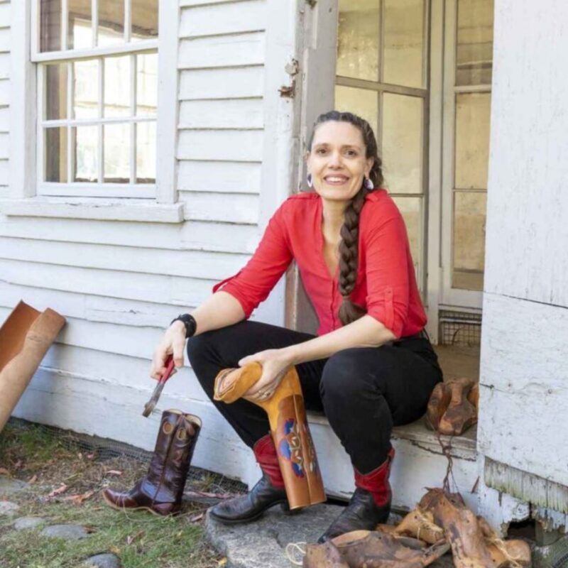A person with a long braid, wearing a red shirt and black pants, sits on the steps of a white wooden building while working on a cowboy boot with a tool. Another boot and pieces of leather are nearby.