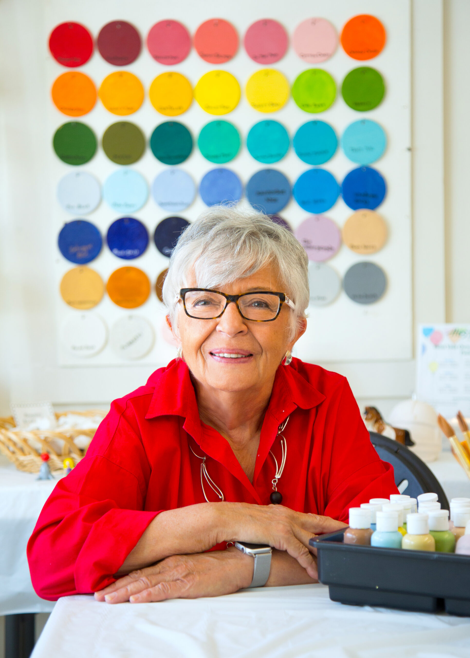 An older woman with short gray hair and glasses sits at a table with Hestia Creations Marblehead handcrafted paint bottles. Behind her, circular color samples are arranged in a grid on the wall. She wears a red shirt and smiles warmly.