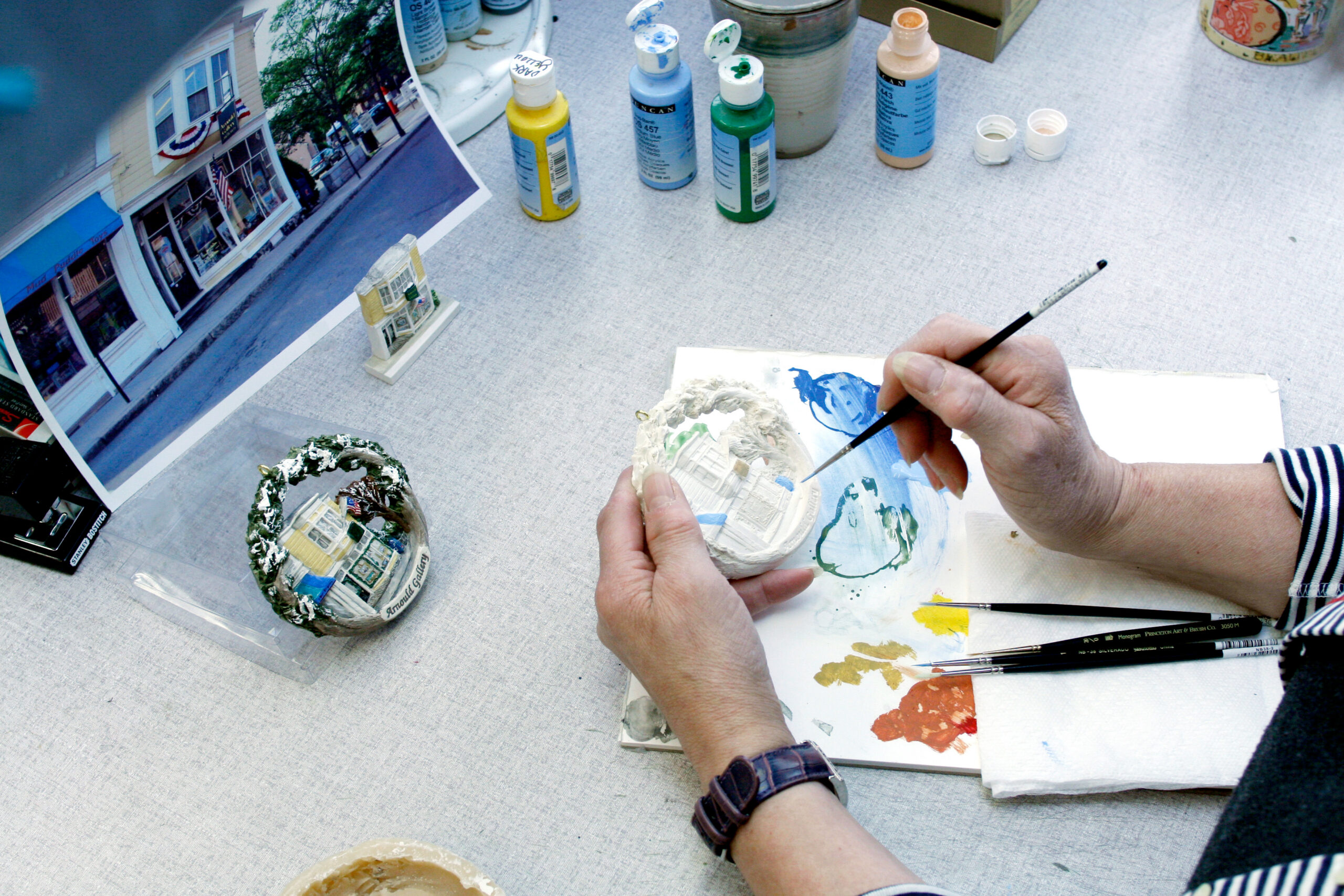 A person paints a handcrafted model building, referencing a printed photo of a Marblehead storefront. Bottles of paint, brushes, and a palette with mixed colors are on the table. The person wears a watch and a striped sleeve, inspired by Hestia Creations.