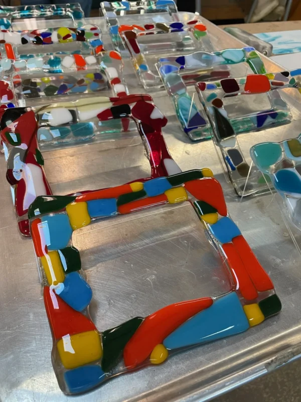 Colorful fused glass picture frames with abstract designs are arranged in rows on a metal tray, featuring a variety of bright colors including red, blue, yellow, green, and white.