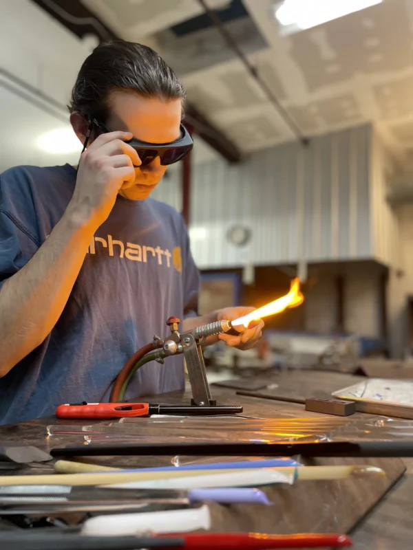 A person wearing protective glasses uses a torch to heat a glass rod at a workbench. Various tools and materials are spread out on the table in front of them in an industrial workshop setting.