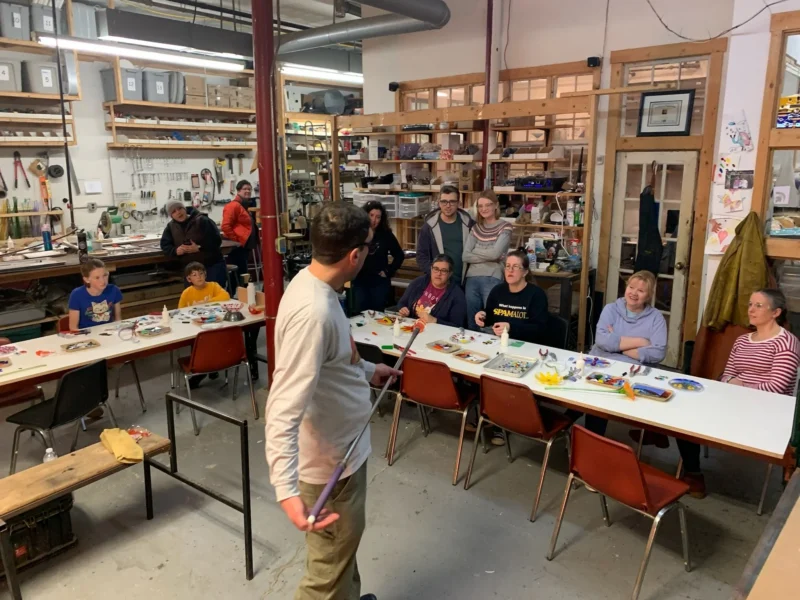 A group of people sit at tables in a workshop space, listening to a person standing at the front. Art supplies and projects are on the tables. Shelves with bins and tools are visible in the background.