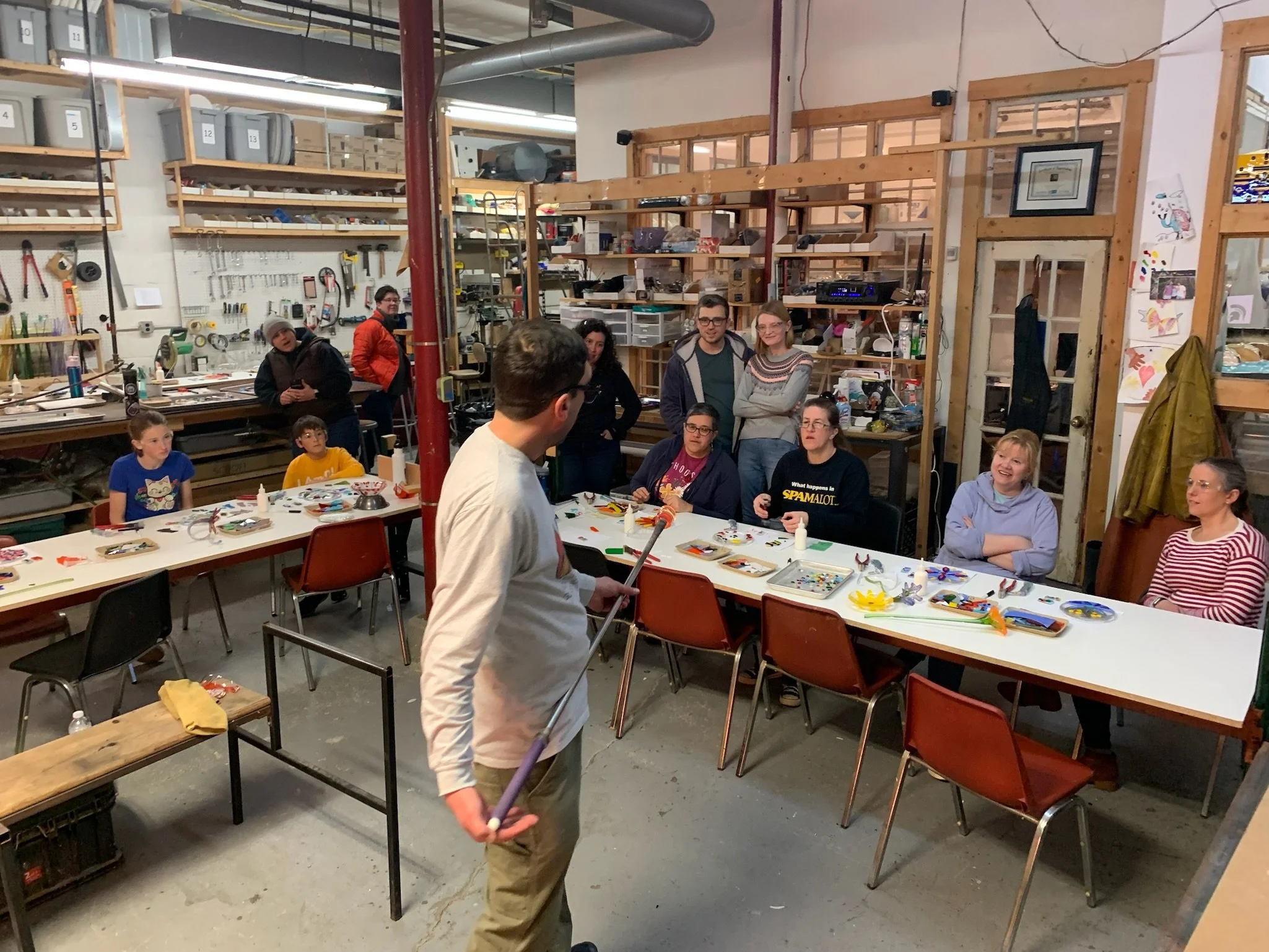 A group of people sit at tables in a workshop space, listening to a person standing at the front. Art supplies and projects are on the tables. Shelves with bins and tools are visible in the background.