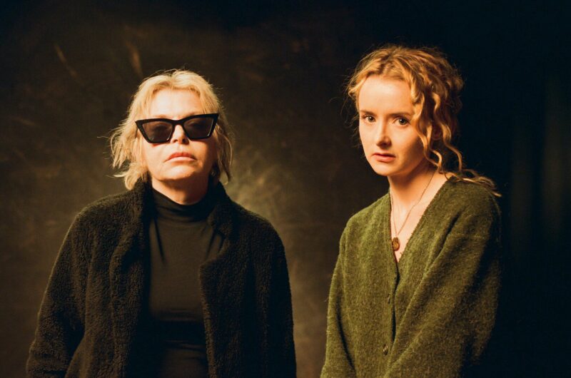 Two women stand side by side in front of a dark background. The woman on the left wears dark sunglasses and a black outfit; the woman on the right has curly hair and wears a green sweater. Both have neutral expressions.