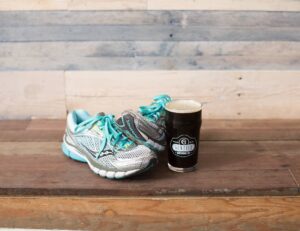 A pair of gray and turquoise running shoes rest on a wooden surface beside a pint glass of dark beer from Gentile Brewing Co., embodying the relaxed spirit of Essex County Sevenings. The backdrop features rustic wood planks.