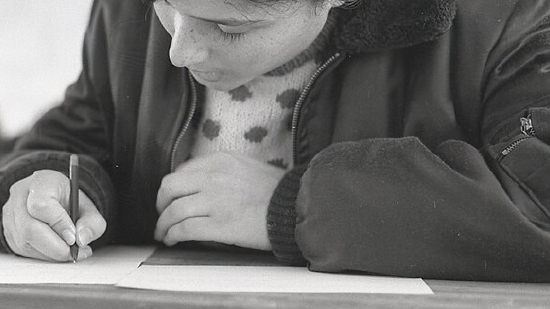 A person wearing a jacket and a polka dot sweater is seated at a table, writing or drawing with a pencil on a sheet of paper. The image is in black and white and taken from an overhead angle.