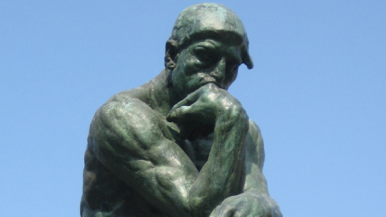 A bronze statue of a muscular man sitting with his chin resting on one hand, appearing deep in thought, set against a clear blue sky.