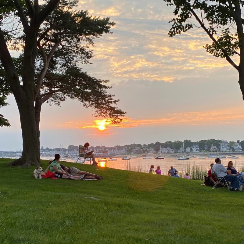 People sit and relax on a grassy area near a waterfront at sunset, with trees framing the scene and boats visible on the water. Some individuals sit in chairs while others sit or lie on the grass with a dog nearby.