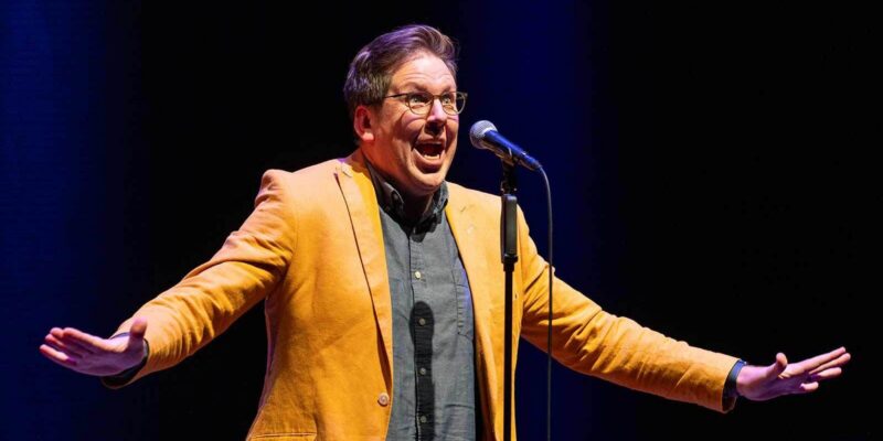 A person wearing glasses, a yellow blazer, and a dark shirt stands on stage in front of a microphone with arms outstretched and an expressive facial expression. The background is dark.