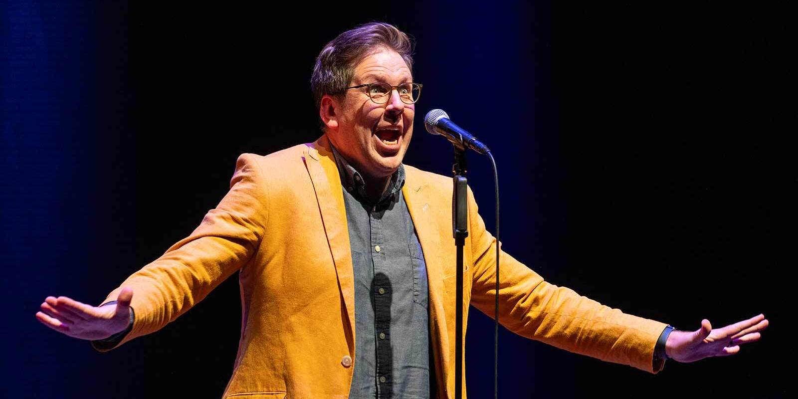 A person wearing glasses, a yellow blazer, and a dark shirt stands on stage in front of a microphone with arms outstretched and an expressive facial expression. The background is dark.