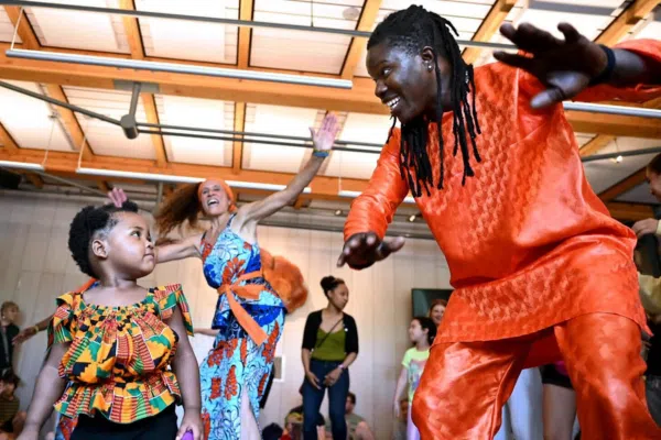 A man in an orange outfit dances enthusiastically with a young girl in a colorful dress at a lively Juneteenth indoor event, while others join the Ammaya Dance and enjoy the rhythmic beat of the drum in the background.