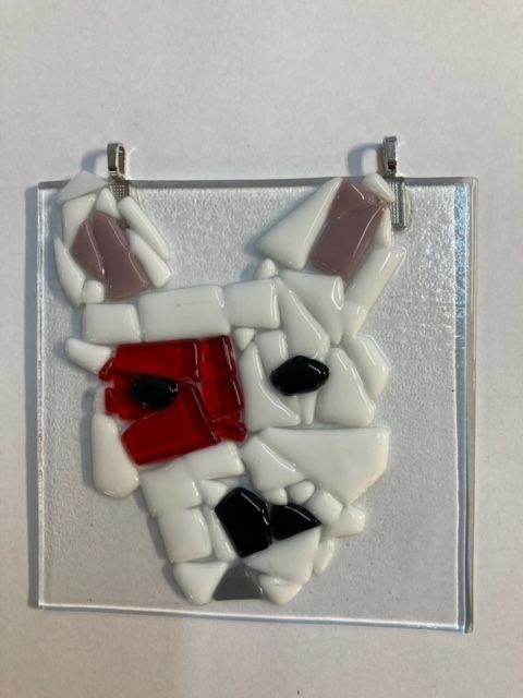 A glass art piece features an abstract white animal face, possibly a dog, made of irregular mosaic pieces with a red patch over one eye—perfect inspiration for our Kids Summer Workshop for ages 10-17. Mounted on a clear square glass panel with metal clips.