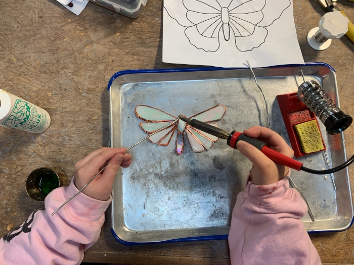 A person in a pink long-sleeve shirt uses a soldering iron to work on a stained glass butterfly at the NSGS Kids Summer Workshop; for ages 10-17, butterfly outlines and tools are set out on the table.