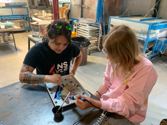 Two people sit at a table in a Kids Summer Workshop, placing colored pieces into a metal tray for a stained glass project. Tools and equipment fill the background, making it perfect for ages 10-17 at NSGS.