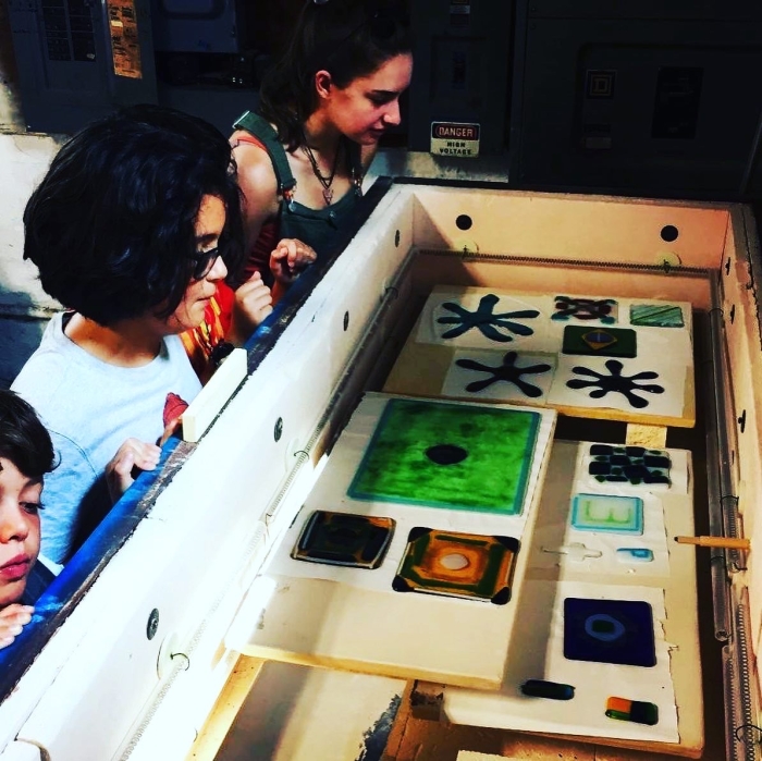 Three people look into a kiln containing fused glass artwork in vibrant shapes and colors, created during the NSGS Junior Summer Workshop for ages 7-10, featuring abstract designs and geometric patterns on square and rectangular tiles.