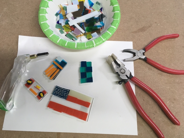 A bowl with colored glass pieces, a glass cutter, pliers, and small rectangular glass mosaics are arranged on white paper atop a brown work surface—perfect for creative hands at the NSGS Junior Summer Workshop for Ages 7-10.