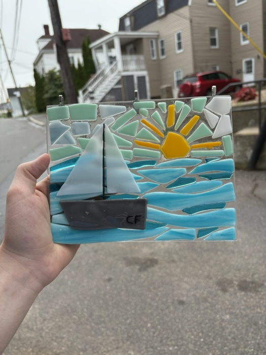 A hand holds up a small glass mosaic from the NSGS Junior Summer Workshop, featuring a sailboat on blue water with a yellow sun rising or setting, and houses and a red car in the background—perfect for ages 7-10.