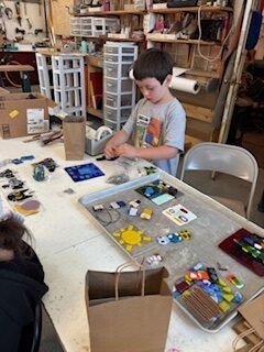 A child, ages 7-10, stands at a work table in a craft studio, assembling colorful glass pieces during the NSGS Junior Summer Workshop. The table is covered with art supplies and finished glass projects. Shelves, tools, and boxes are visible in the background.