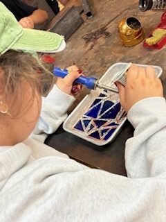 At the NSGS Junior Summer Workshop for Ages 7-10, a child in a gray sweatshirt uses a soldering tool to assemble a geometric stained glass design on a tray, with art supplies scattered on the wooden table and a green towel draped over their head.