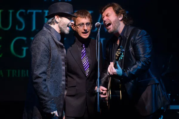Three men in suits, resembling the Australian Bee Gees, stand close together singing into a shared microphone on stage; one holds an acoustic guitar, and another wears a hat and glasses. Text is partially visible in the background.