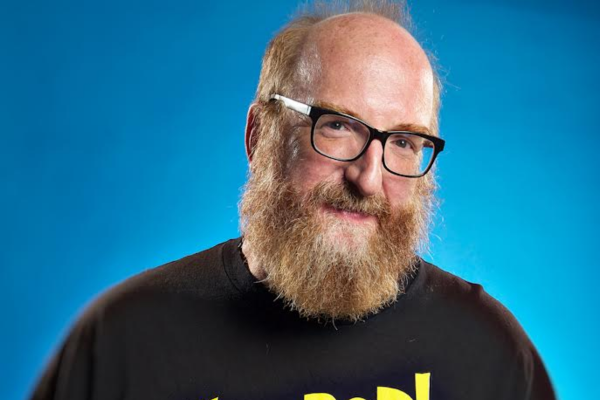 A man with a full reddish-brown beard and glasses is smiling slightly. He is wearing a black T-shirt with yellow text. The background is a solid blue gradient.