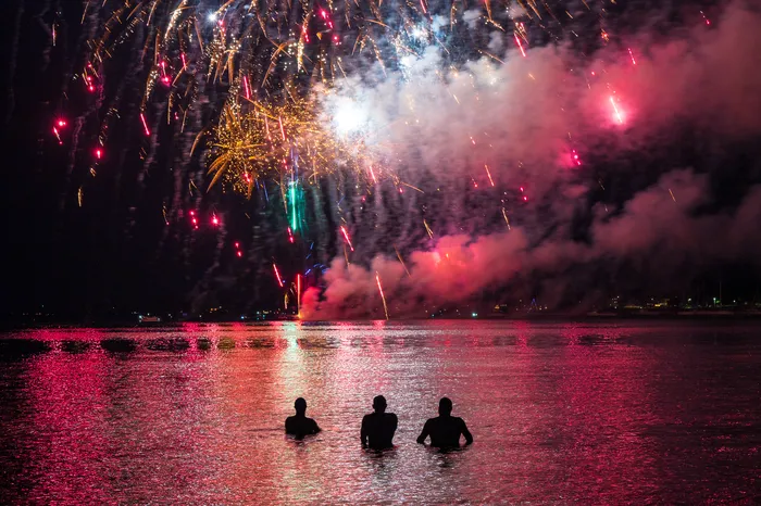 Three people stand waist-deep in water at night, watching colorful fireworks explode in the sky above, reflecting red and yellow light on the surface of the water.