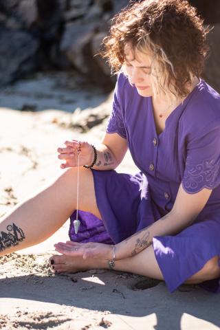 A person with curly hair wearing a purple dress sits on the sand, holding a pendulum over their hand. They have several tattoos on their arms and leg, and appear focused on the pendulum's movement.