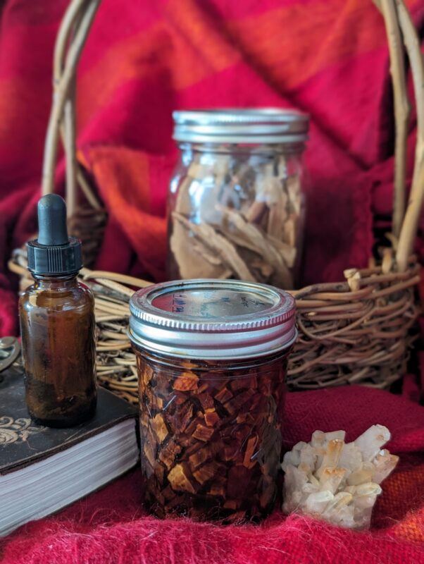 A glass jar filled with dried herbs, a brown dropper bottle, a jar with wood pieces, a cluster of crystals, and a wicker basket are arranged on a red fabric background.