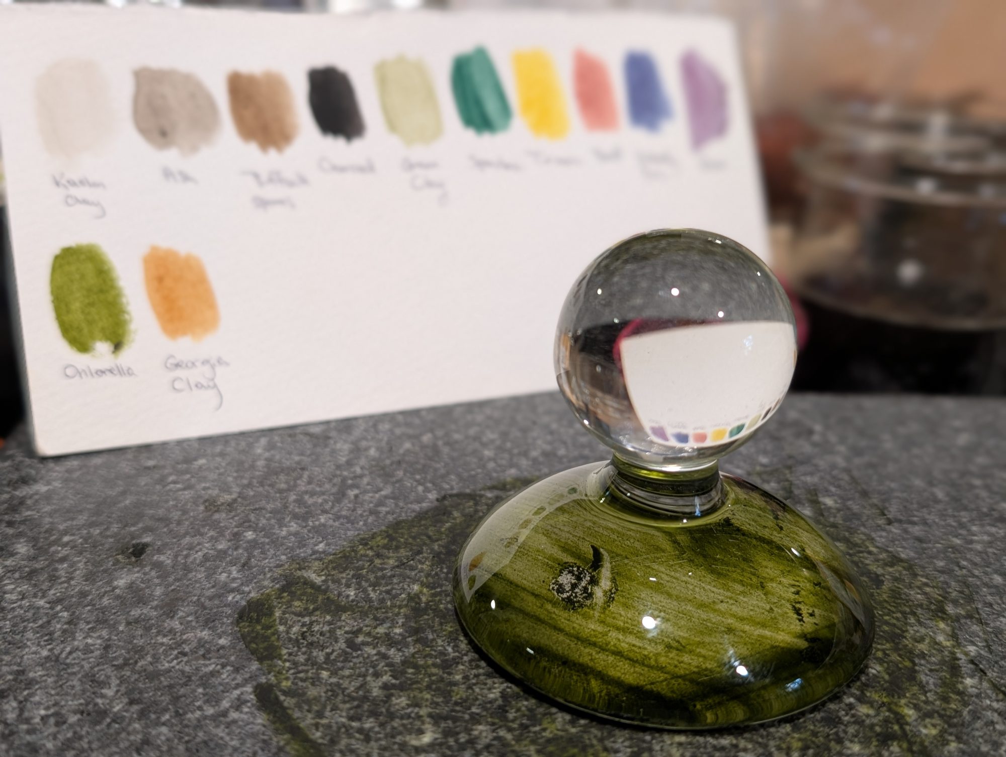 A glass ink holder with green ink sits on a gray surface. In the background, a card displays swatches of various colors labeled with names. Two glass jars are partially visible on the right.