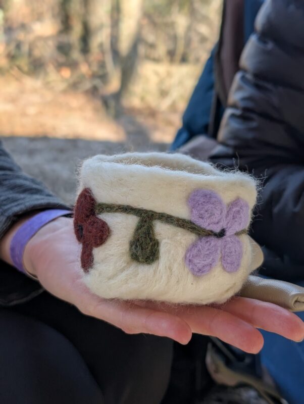 A person holds a small, cream-colored felted cup decorated with purple and red flower designs. The background shows another person in a puffy jacket and an outdoor setting with blurred trees.