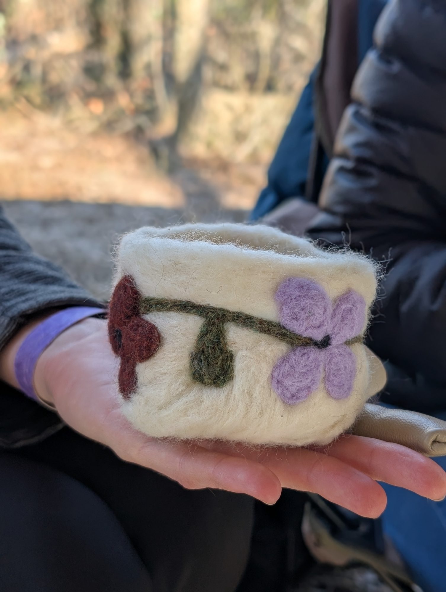 A person holds a small, cream-colored felted cup decorated with purple and red flower designs. The background shows another person in a puffy jacket and an outdoor setting with blurred trees.