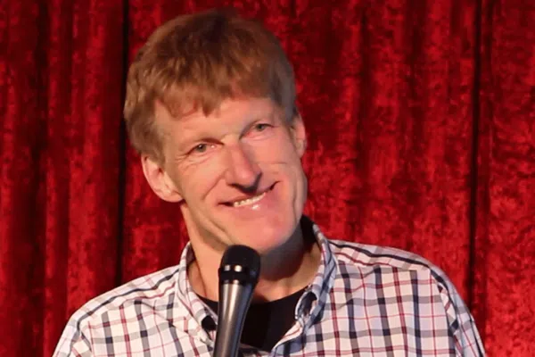 A man with short, light brown hair is smiling while holding a microphone. Don McMillan is wearing a white and blue plaid shirt and stands in front of a red curtain.