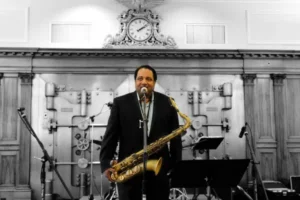 A man in a suit stands in front of a microphone with a saxophone, ready to perform at an Essex County CCE Nightlife event. Behind him is a large metal vault door and clock above wood paneling, with musical instruments and equipment surrounding him.