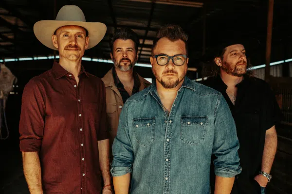 Four men stand side by side indoors. One, from the Eli Young Band, sports a cowboy hat and maroon shirt, another wears glasses and a denim shirt, while the remaining two wear dark shirts against a dimly lit background.