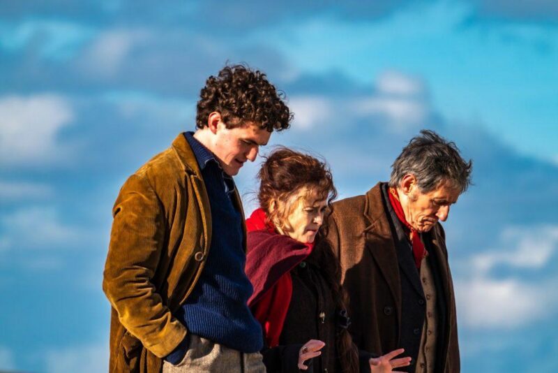 Three people are walking outdoors under a blue sky with clouds. The younger man on the left has curly hair and wears a brown coat. The older woman in the middle and older man on the right are talking while walking closely together.