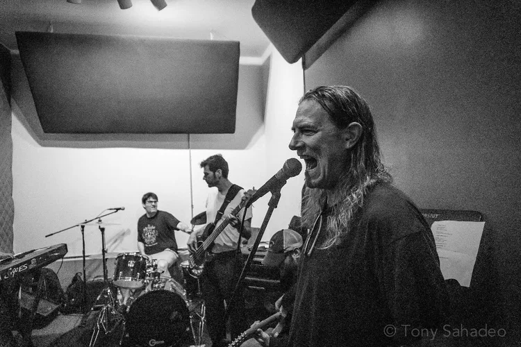 Four musicians rehearse in a studio at Drink n Draw. One person sings passionately into a microphone in the foreground, while others play drums, guitar, and keyboard in the background. The photo is in black and white.