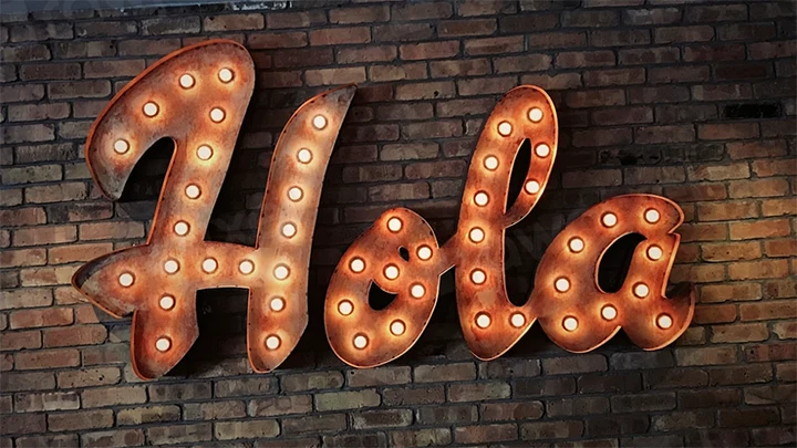 A large metal sign spelling "Hola" is mounted on a brick wall. The sign is lit with multiple round light bulbs outlining each letter.