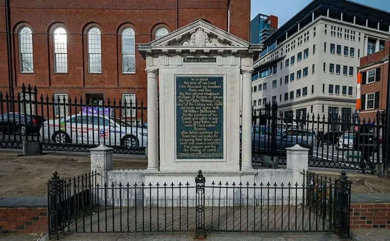A stone monument with an inscription stands behind a black iron fence on the sidewalk—an intriguing sight often featured on the Boston Witchcraft Tour. Brick and modern buildings rise in the background, with cars parked along the street.