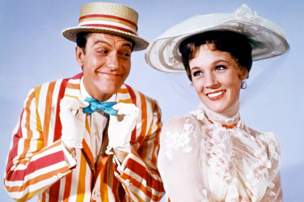 A man in a striped jacket, bow tie, and straw boater hat stands next to a woman in a high-necked, lacy white dress and wide-brimmed hat against a plain background. Both are smiling.