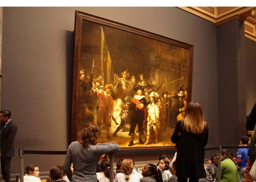 A group of people, some seated and some standing, observe a large framed painting displayed on a museum wall. The painting depicts a historical scene with several figures.