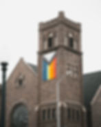 A stone church building with a square clock tower displays a large Progress Pride flag on the front, featuring rainbow and chevron colors. The sky is overcast.